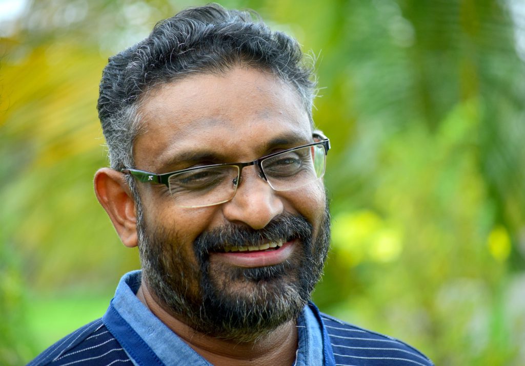 Benyamin with a greying beard and glasses, smiling, in a blue shirt against green foliage