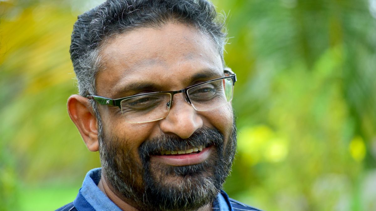 Benyamin with a greying beard and glasses, smiling, in a blue shirt against green foliage