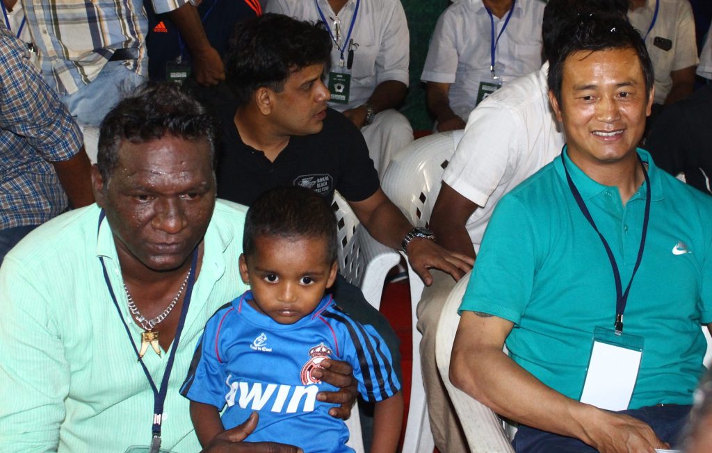 Bhaichung Bhutia smiling in a green polo shirt at a stadium event, with I. M. Vijayan beside him