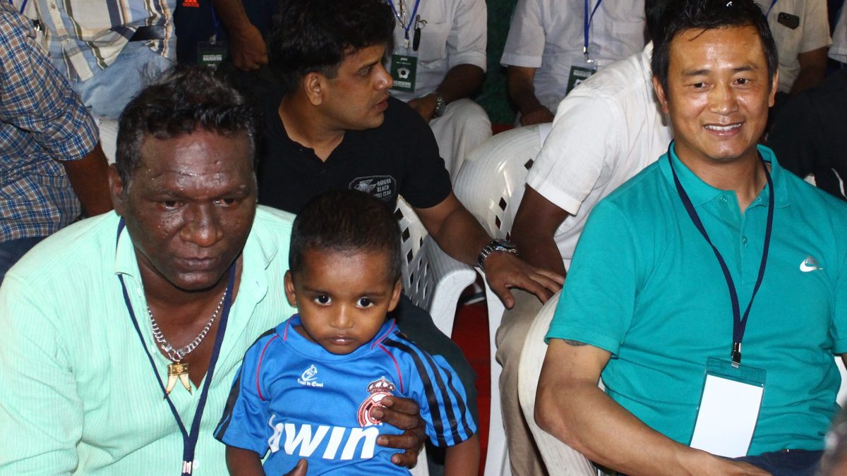 Bhaichung Bhutia smiling in a green polo shirt at a stadium event, with I. M. Vijayan beside him