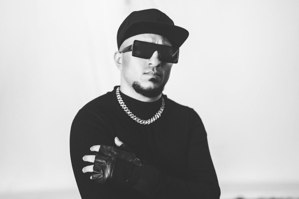 Black and white photo of Brodha V in a cap, sunglasses and chain with his arms crossed