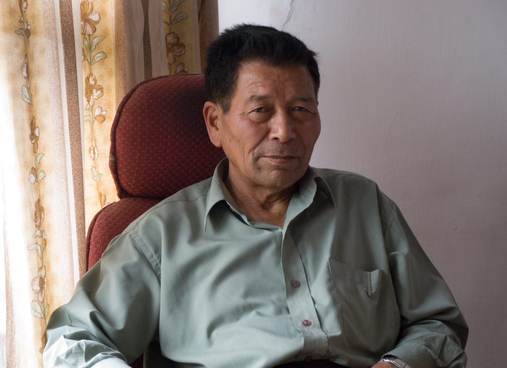 Chewang Norphel in a green shirt seated in an armchair by a window