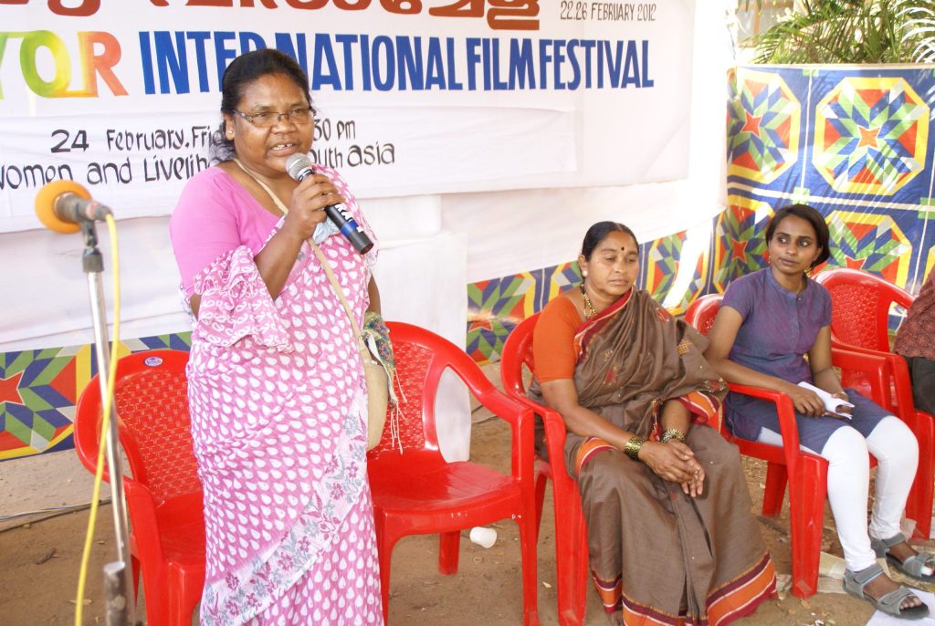 Dayamani Barla in a pink sari speaking into a microphone, with three women seated beside her