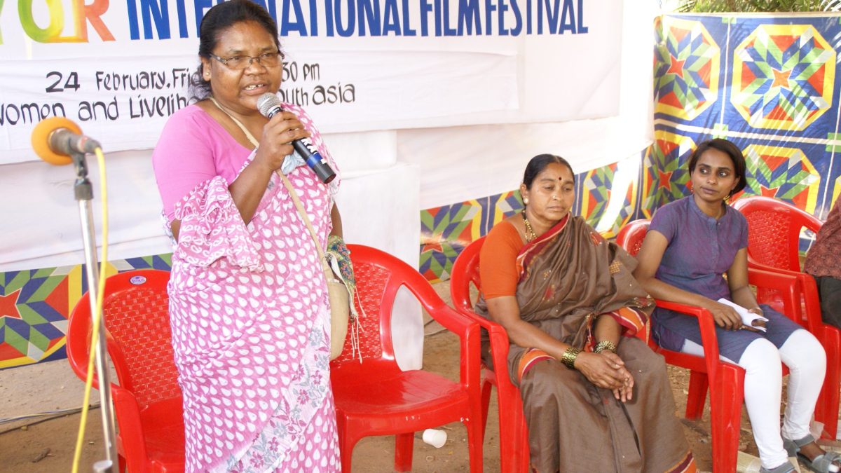 Dayamani Barla in a pink sari speaking into a microphone, with three women seated beside her
