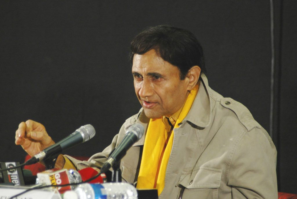 Dev Anand in a khaki jacket and yellow scarf speaking at press microphones