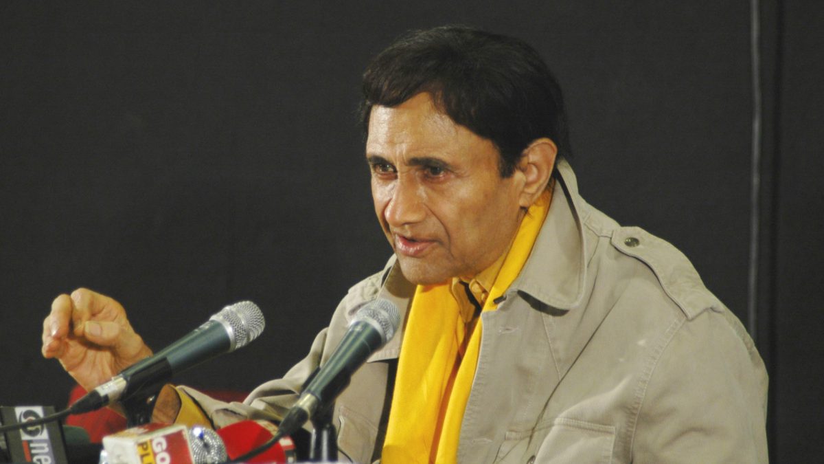 Dev Anand in a khaki jacket and yellow scarf speaking at press microphones