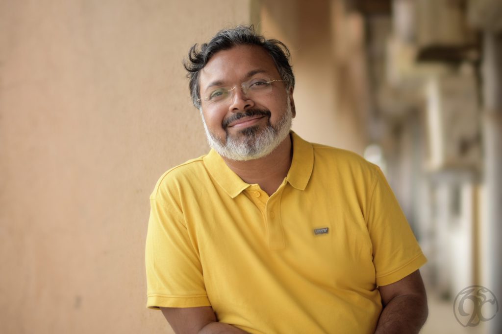 Devdutt Pattanaik with a grey beard and glasses, smiling in a yellow polo shirt