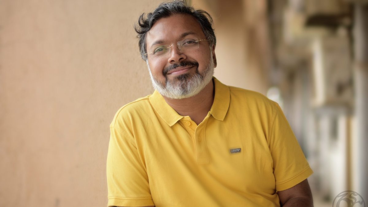 Devdutt Pattanaik with a grey beard and glasses, smiling in a yellow polo shirt