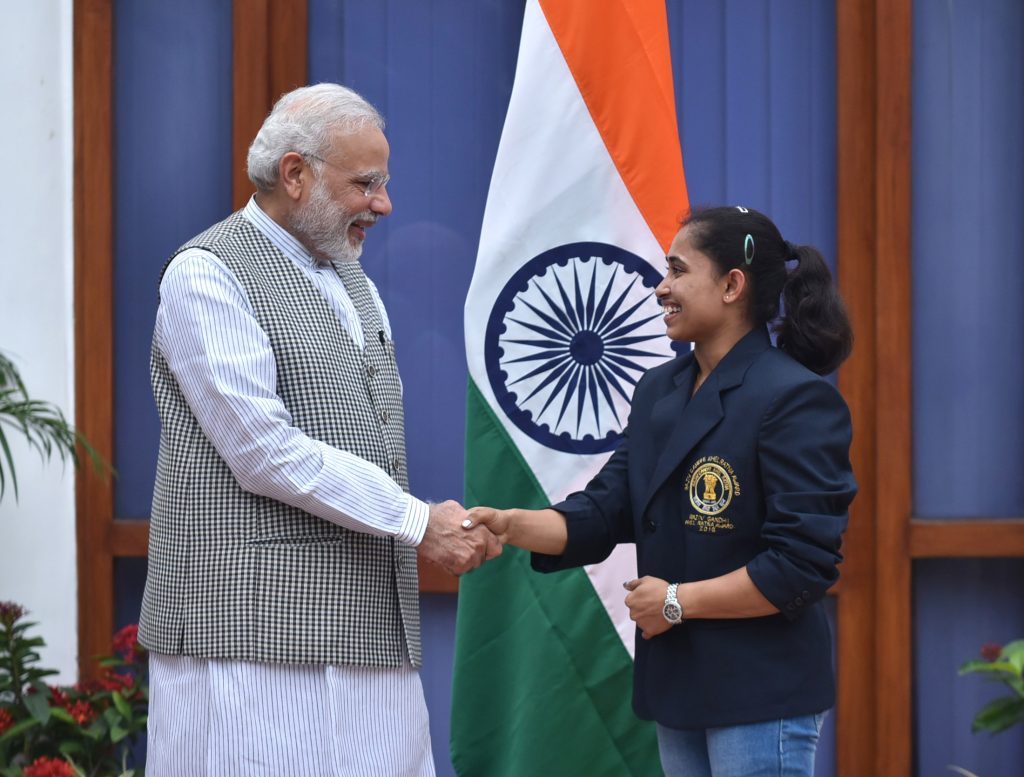 Dipa Karmakar in a navy blazer shaking hands with Prime Minister Narendra Modi beside the Indian flag