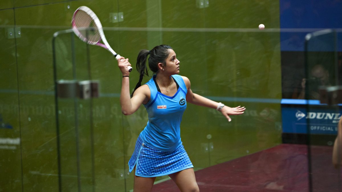 Dipika Pallikal in a blue dress about to hit a squash ball on a glass court