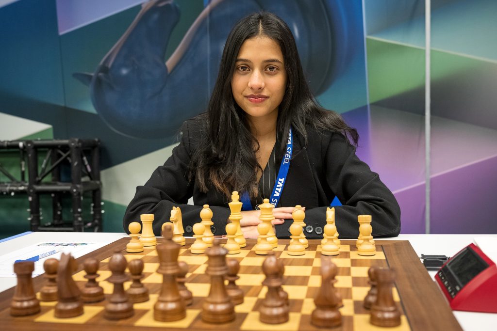 Divya Deshmukh in a black jacket seated behind a chessboard, looking at the camera