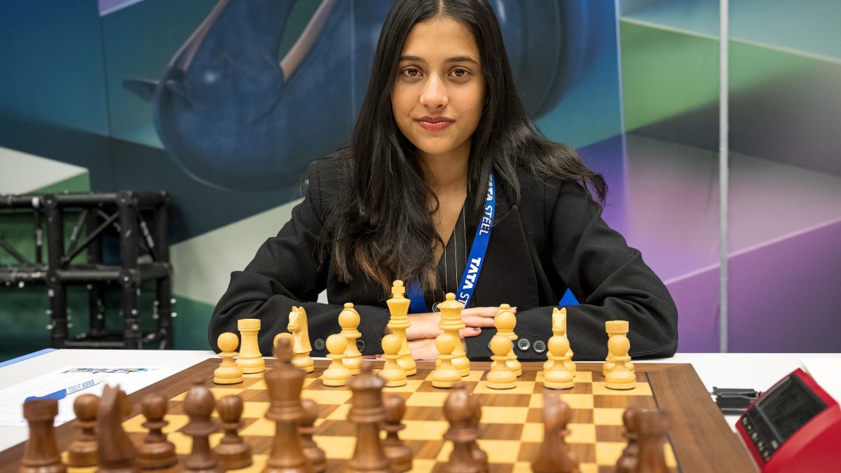 Divya Deshmukh in a black jacket seated behind a chessboard, looking at the camera