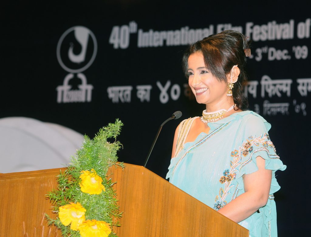 Divya Dutta in a light blue sari speaking at a lectern decorated with flowers