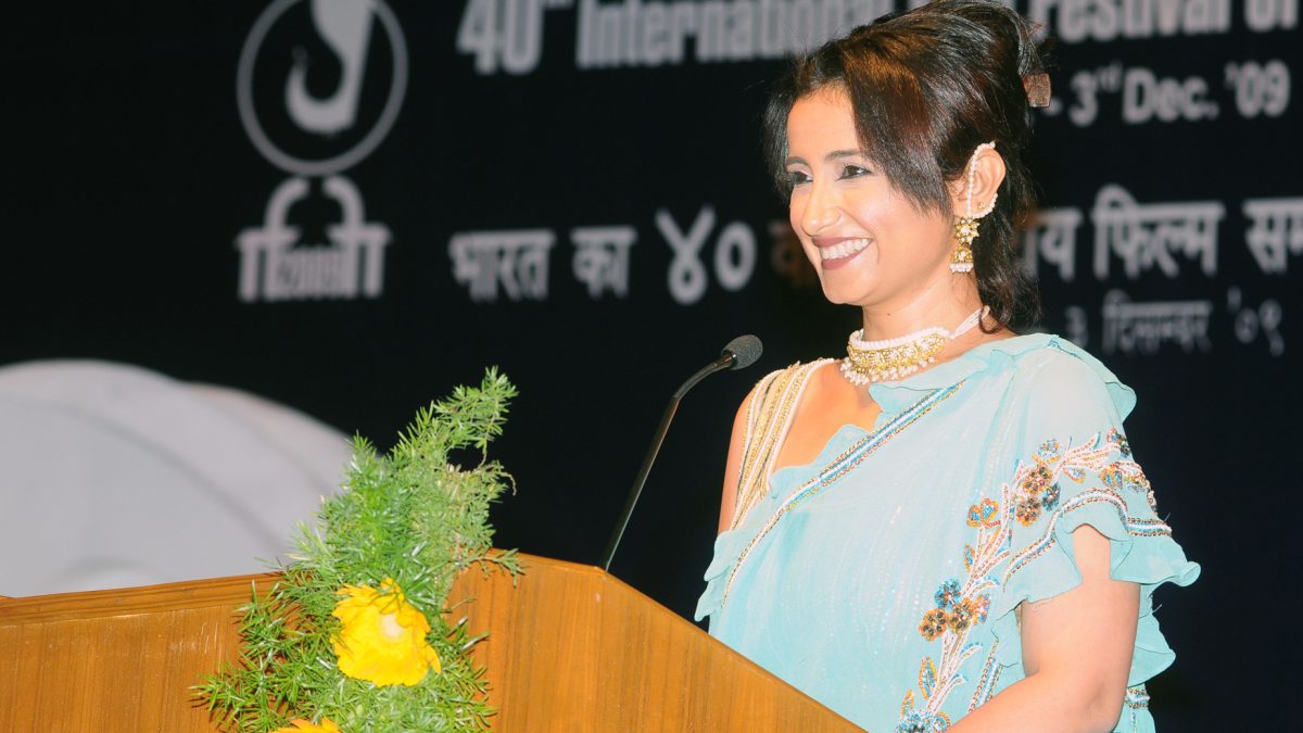 Divya Dutta in a light blue sari speaking at a lectern decorated with flowers