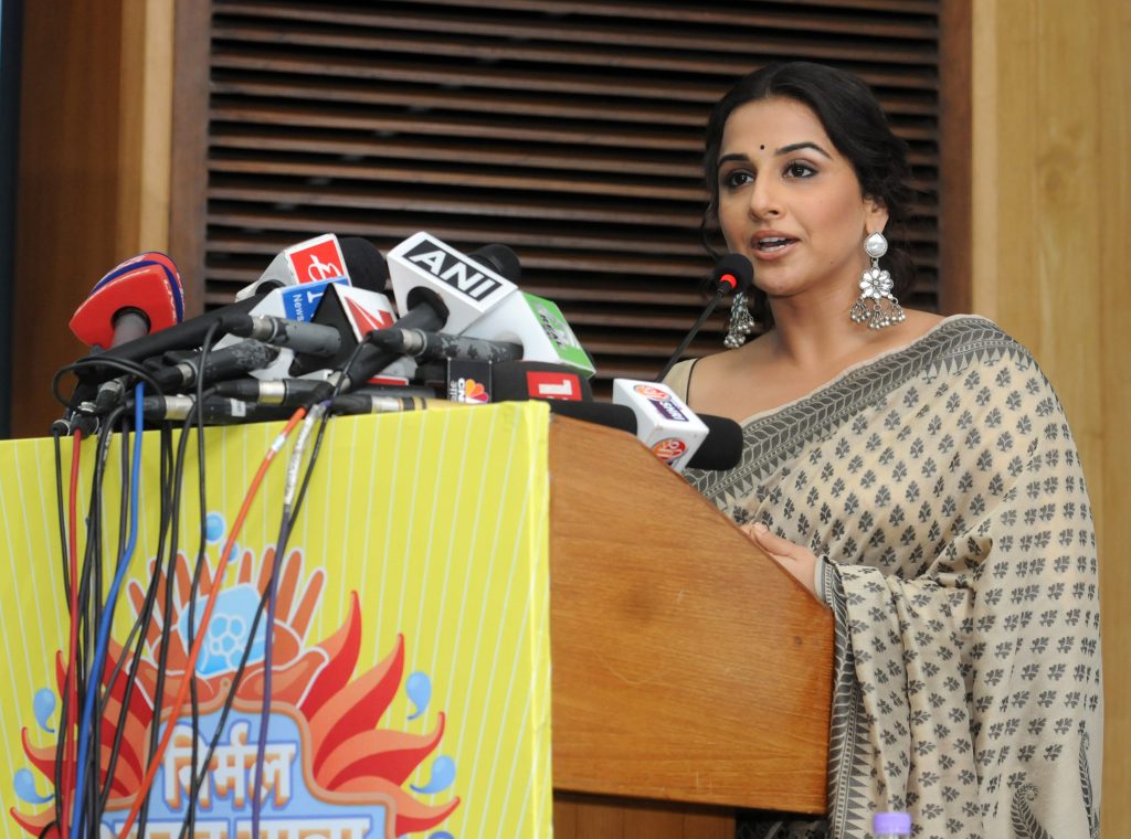 Actress Vidya Balan speaking at a podium crowded with microphones