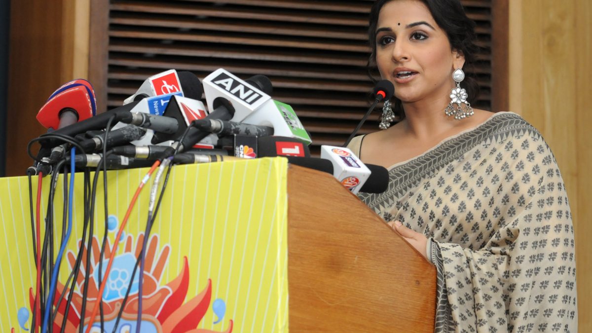 Actress Vidya Balan speaking at a podium crowded with microphones
