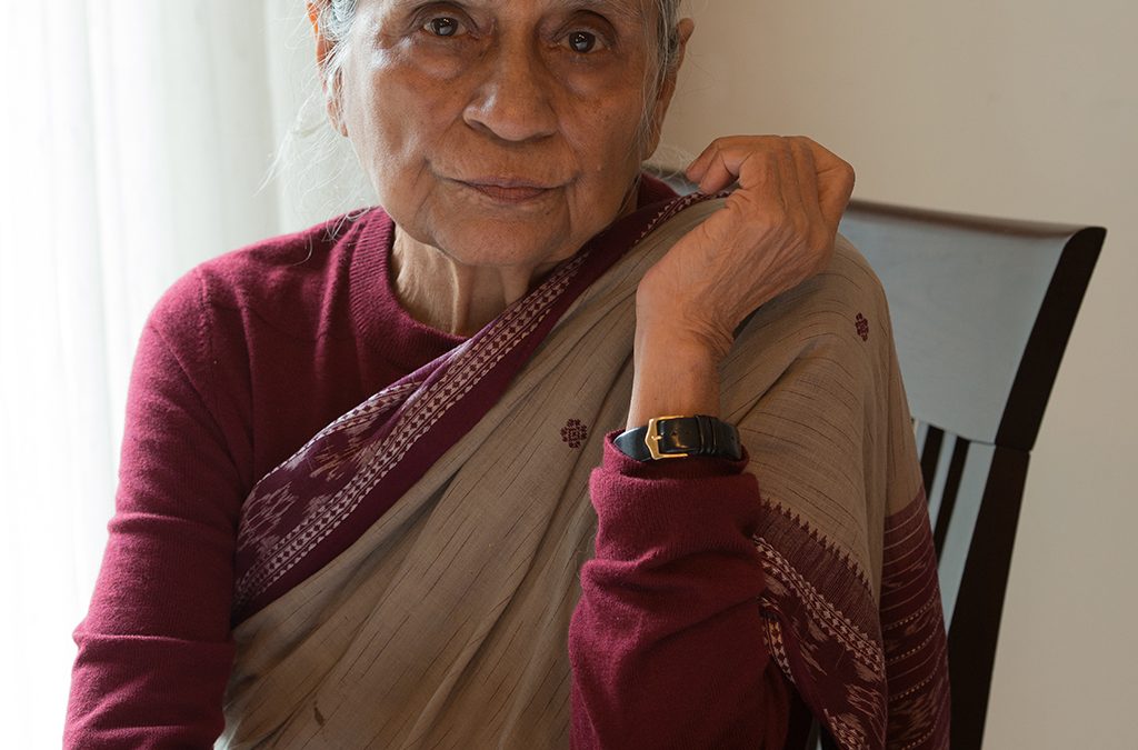 Ela Bhatt in a maroon blouse and brown sari seated at a table, resting her chin on her hand