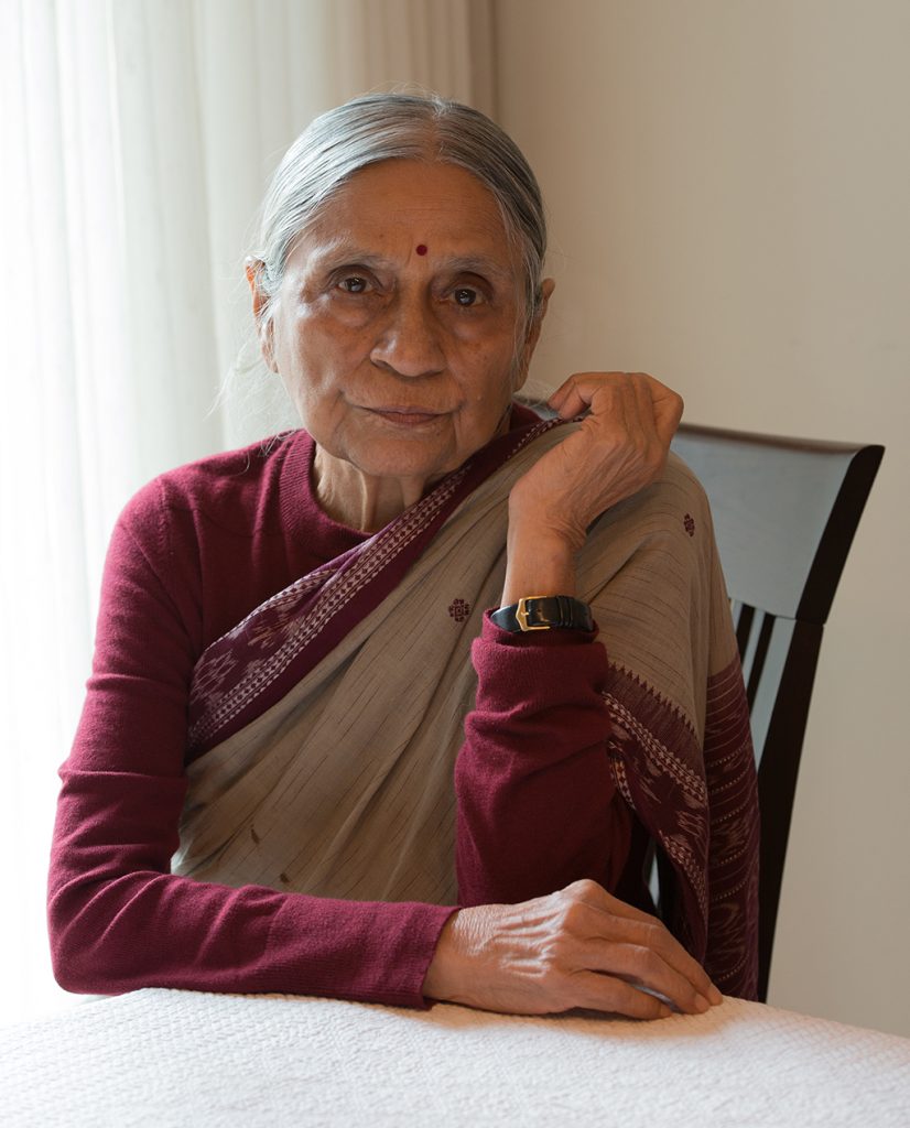 Ela Bhatt in a maroon blouse and brown sari seated at a table, resting her chin on her hand