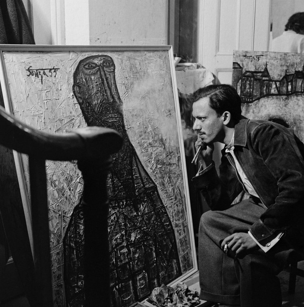 A black-and-white photograph of F. N. Souza seated before one of his paintings