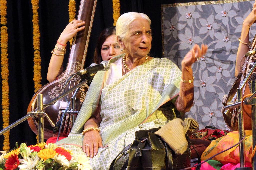 Girija Devi singing on stage, seated among tanpura players and flowers