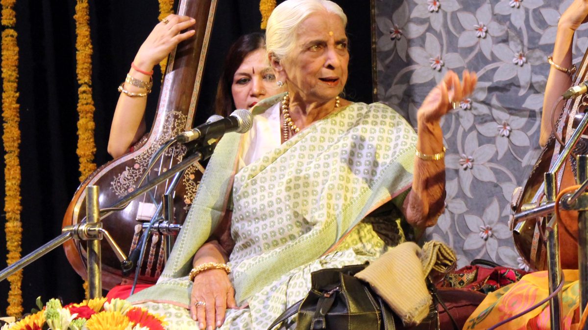 Girija Devi singing on stage, seated among tanpura players and flowers