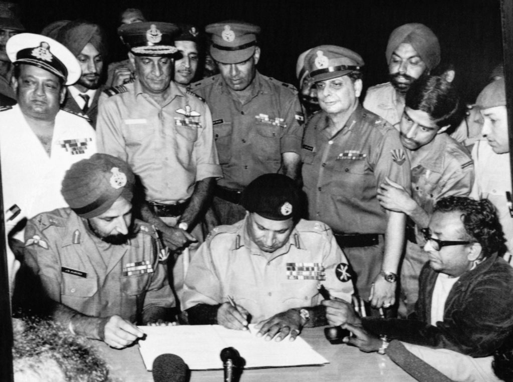 Lt Gen A. A. K. Niazi signing the Instrument of Surrender beside Lt Gen Jagjit Singh Aurora at Dhaka on 16 December 1971, with Indian officers standing behind