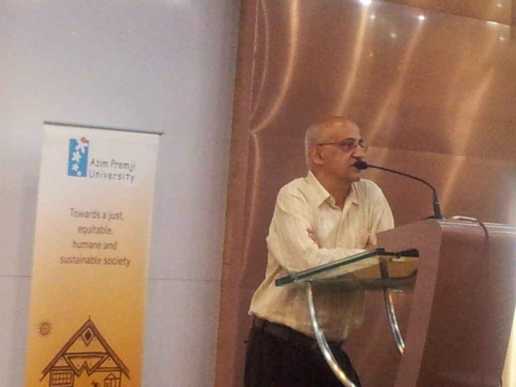 Harsh Mander in a cream shirt speaking at a glass lectern beside an Azim Premji University banner
