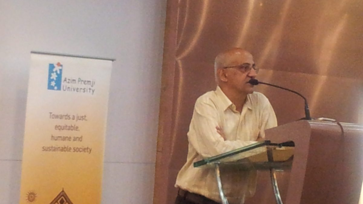 Harsh Mander in a cream shirt speaking at a glass lectern beside an Azim Premji University banner
