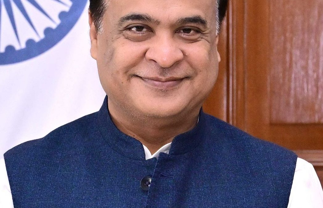 Himanta Biswa Sarma in glasses and a blue waistcoat, beside the national flag