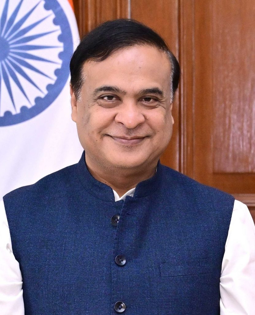Himanta Biswa Sarma in glasses and a blue waistcoat, beside the national flag