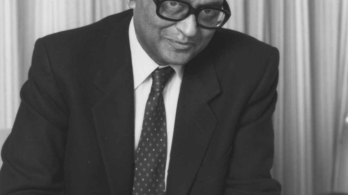 A black-and-white photograph of I. G. Patel in glasses, writing at a desk