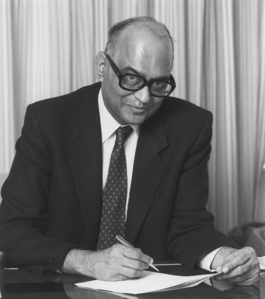 A black-and-white photograph of I. G. Patel in glasses, writing at a desk