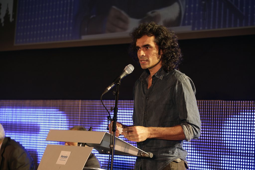 Imtiaz Ali with long curly hair speaking at a lectern