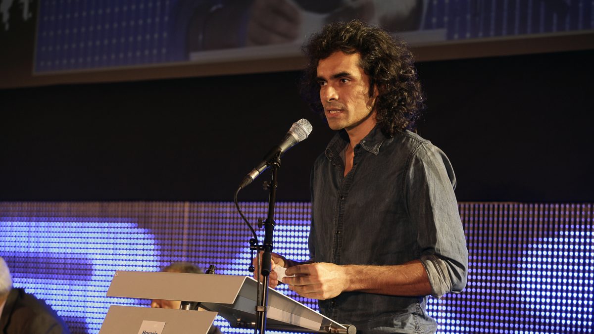 Imtiaz Ali with long curly hair speaking at a lectern