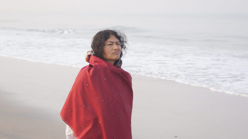 Irom Sharmila wrapped in a red shawl standing on a beach