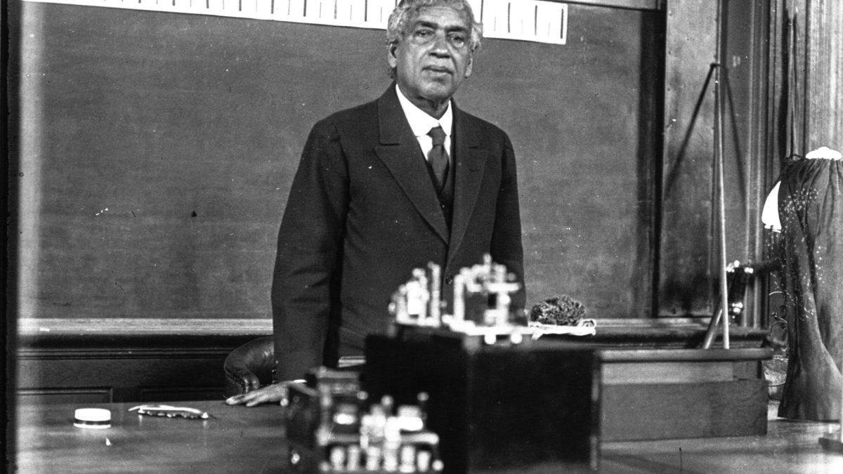 Jagadish Chandra Bose standing behind a table of instruments in a lecture hall, with a blackboard behind him