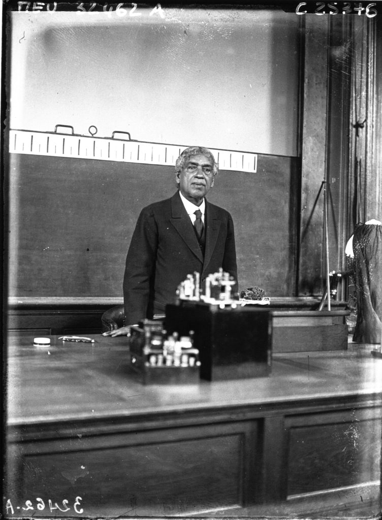 Jagadish Chandra Bose standing behind a table of instruments in a lecture hall, with a blackboard behind him