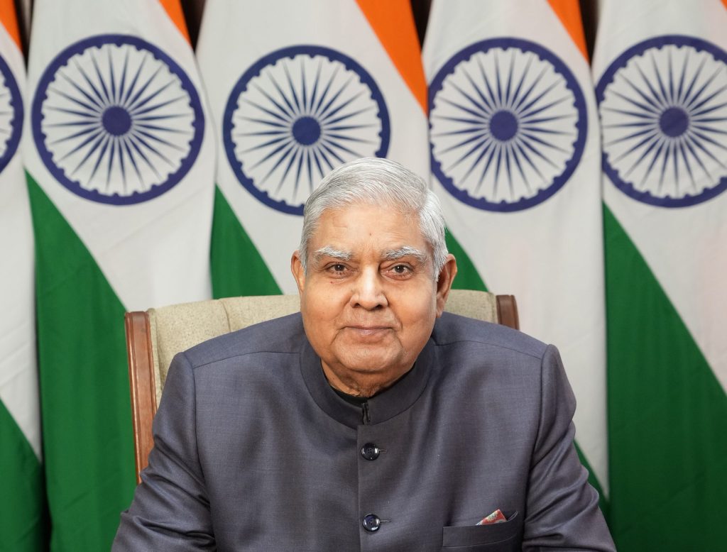 Jagdeep Dhankhar in a grey bandhgala seated in front of Indian national flags