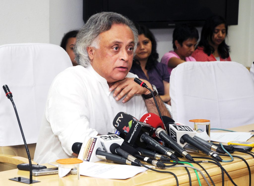 Jairam Ramesh with white hair in a white kurta speaking at press microphones