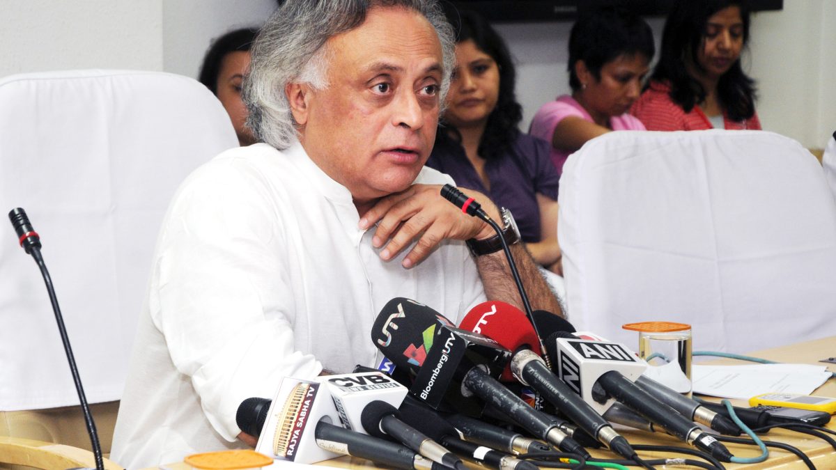 Jairam Ramesh with white hair in a white kurta speaking at press microphones