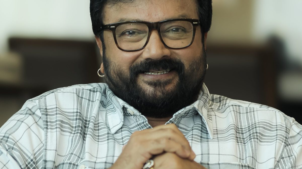 Jayaram with a beard and glasses, in a checked shirt, hands clasped