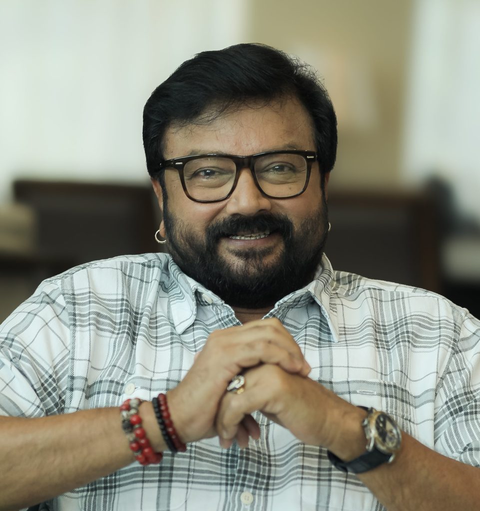 Jayaram with a beard and glasses, in a checked shirt, hands clasped
