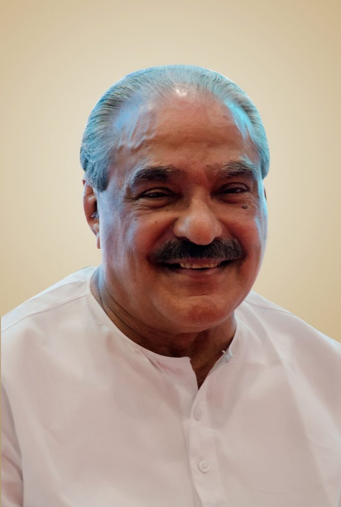 K. M. Mani with white hair and a moustache, smiling, in a white shirt