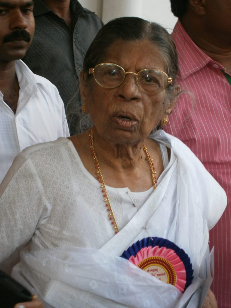 K. R. Gouri Amma in glasses and a white sari, wearing a rosette