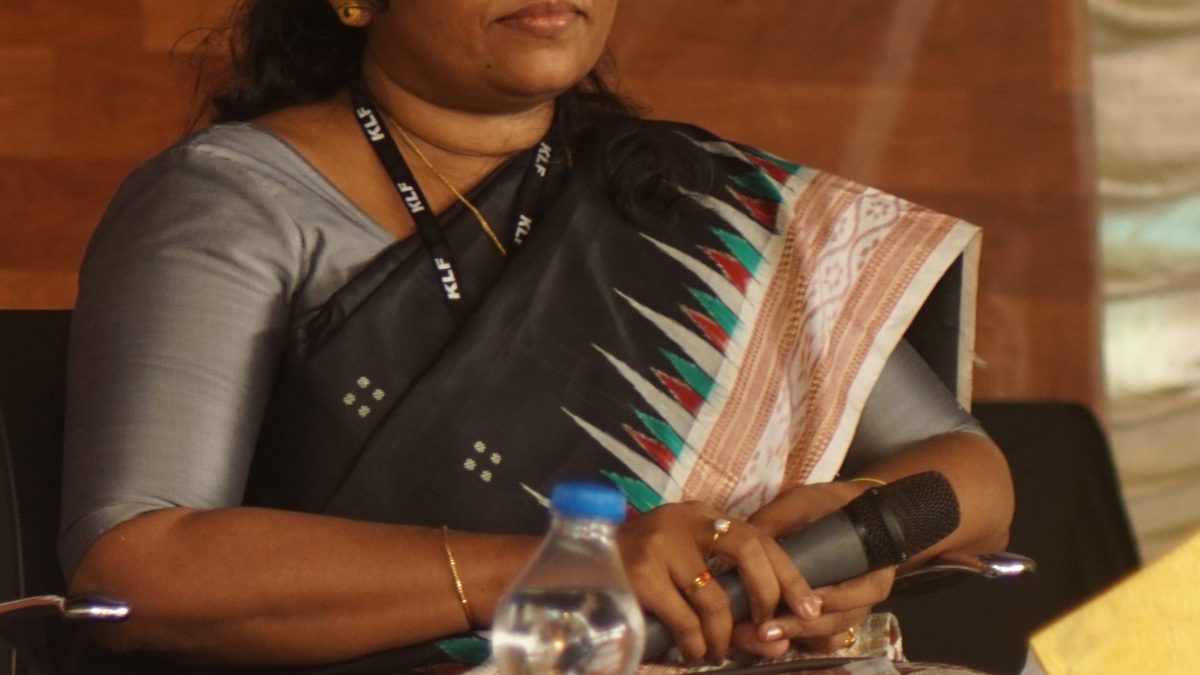 K. R. Meera in a grey sari seated on stage with a microphone