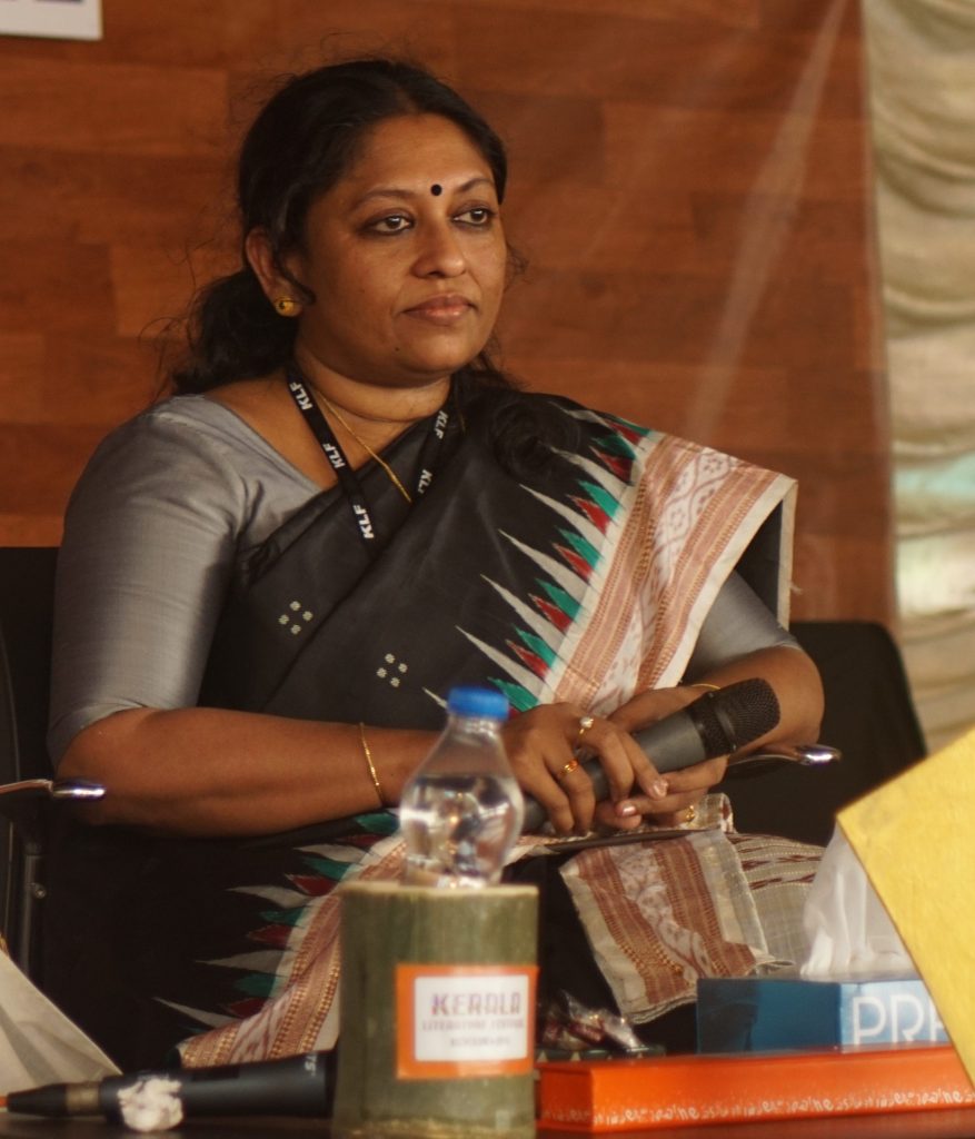 K. R. Meera in a grey sari seated on stage with a microphone