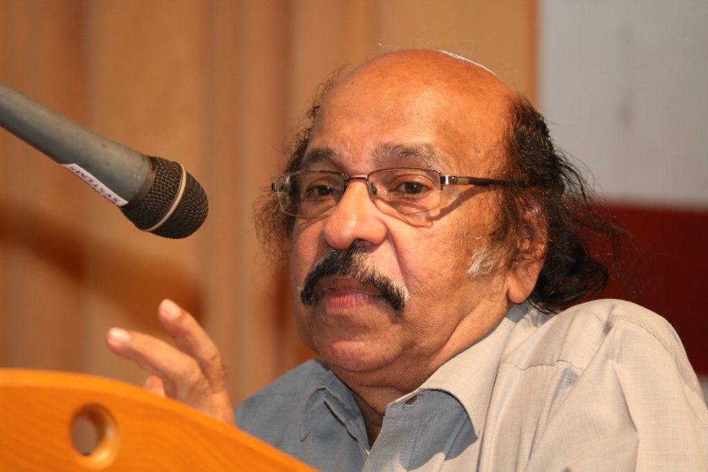 K. Satchidanandan with a moustache and glasses, speaking at a microphone