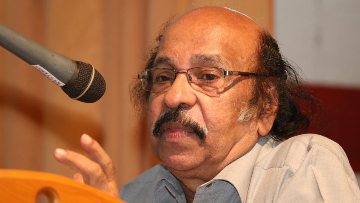 K. Satchidanandan with a moustache and glasses, speaking at a microphone