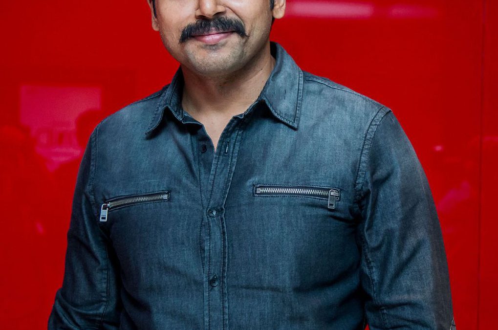 Karthi with a moustache, in a denim shirt, against a red backdrop