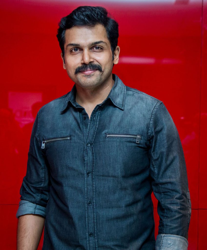 Karthi with a moustache, in a denim shirt, against a red backdrop